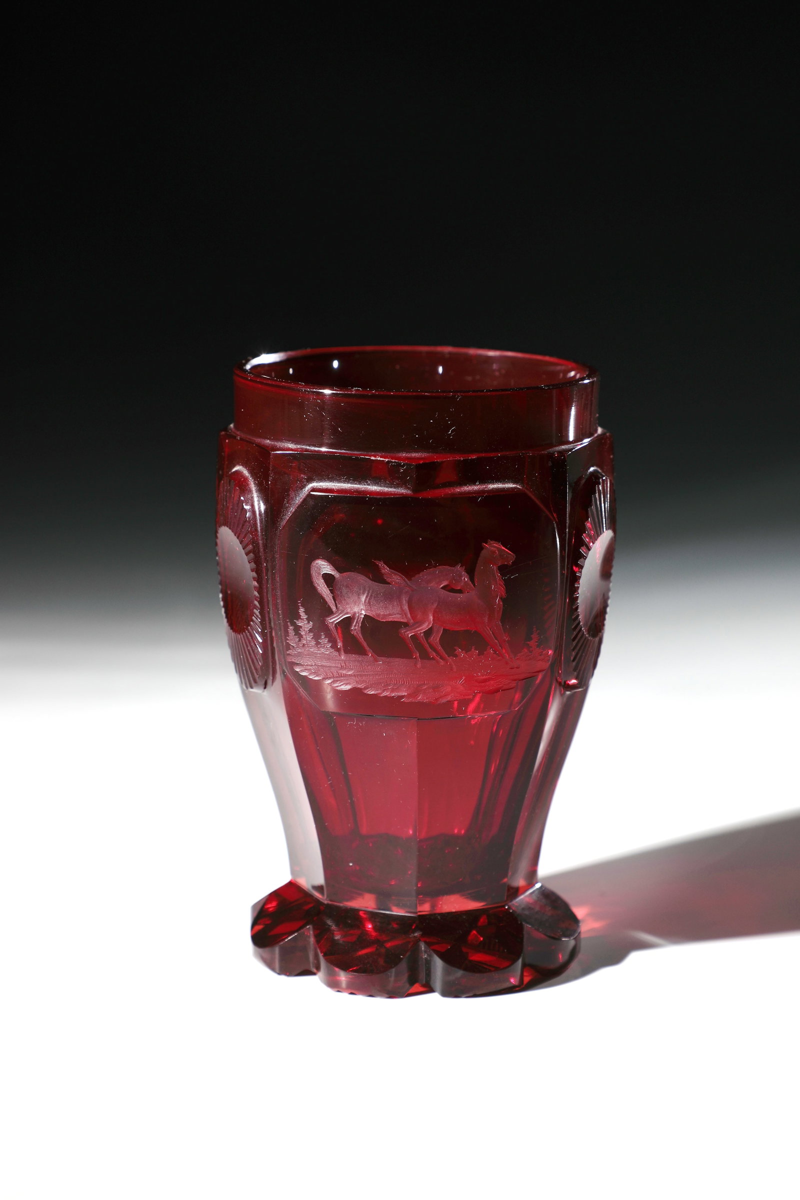 Mug with horses: Beaker with horses Bohemia, 2nd half of the 19th century Colourless, ruby-red glazed glass. Hatched floor. Passively sharpened foot. Eight-fold peeled body with six high-cut rosettes. Frontal