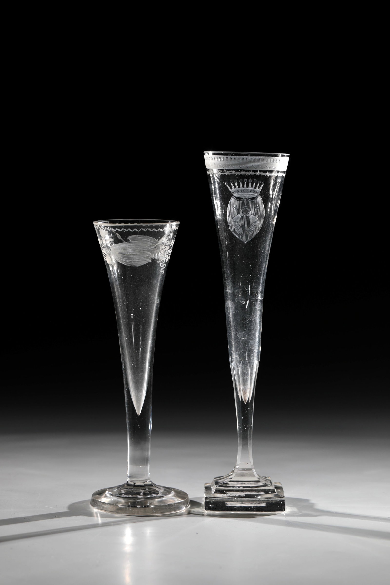 Two champagne flutes: Two champagne flutes Bohemia, 18th century Colourless glass. Square or round, graduated plinth. Trumpet-shaped cuppa with cut borders below the lip rim. One with a coat of arms. H. 17,5 - 22 cm Zwei
