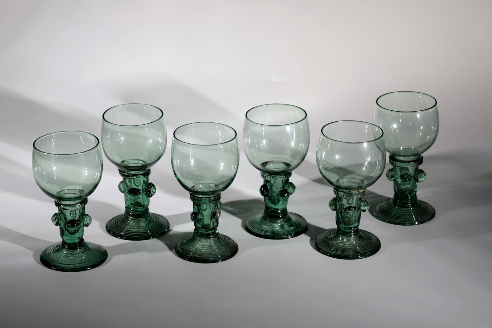 Six Romans: Six Romans German, 18th century Green glass with tear-off. Slightly rising, spun foot. Hollow shaft open at the top with four applied berry nubs. Thread support pinched at the base of the bulbous