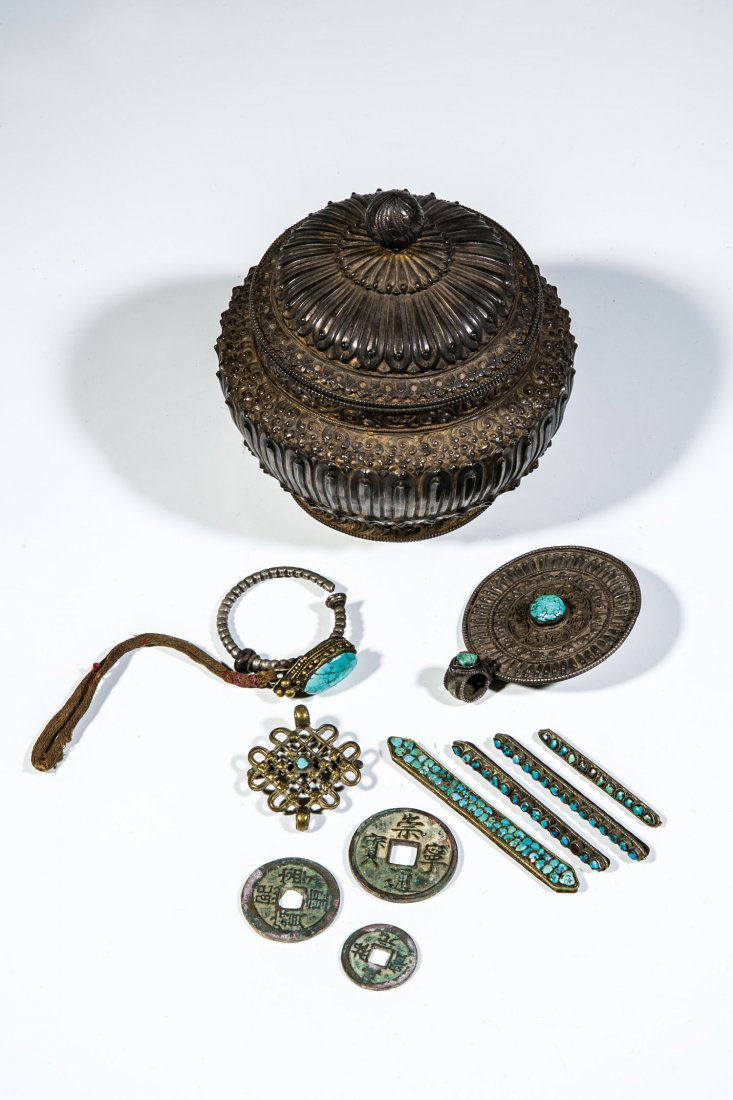 Deckelgefäß mit 10 Münzen und 6 Schmuckteilen: Deckelgefäß mit 10 Münzen und 6 Schmuckteilen Tibet, 19./20. Jh. Weißmetall. Korpus des Deckelgefäßes umlaufend im Flachrelief mit stilisierten Lotosblättern sowie Schnörkel-Ornamenten gearbei