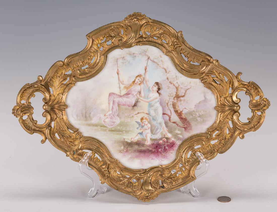 French Porcelain & Ormolu Plaque: French Sevres style shaped porcelain plaque depicting two young women in an outdoor setting, one on a swing, with a cherub at their feet, signed lower left "Miguel" and housed in a Rococo style gilt b