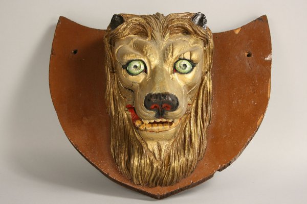Circus Carnival Plaque, Lion's Head: Early circus carnival wooden plaque in shield design mounted with carved lion head painted in gold, green, red and black. Condition - Age cracks with later 20th repaint. 12 3/4" H x 14 1/2" W. Late 19