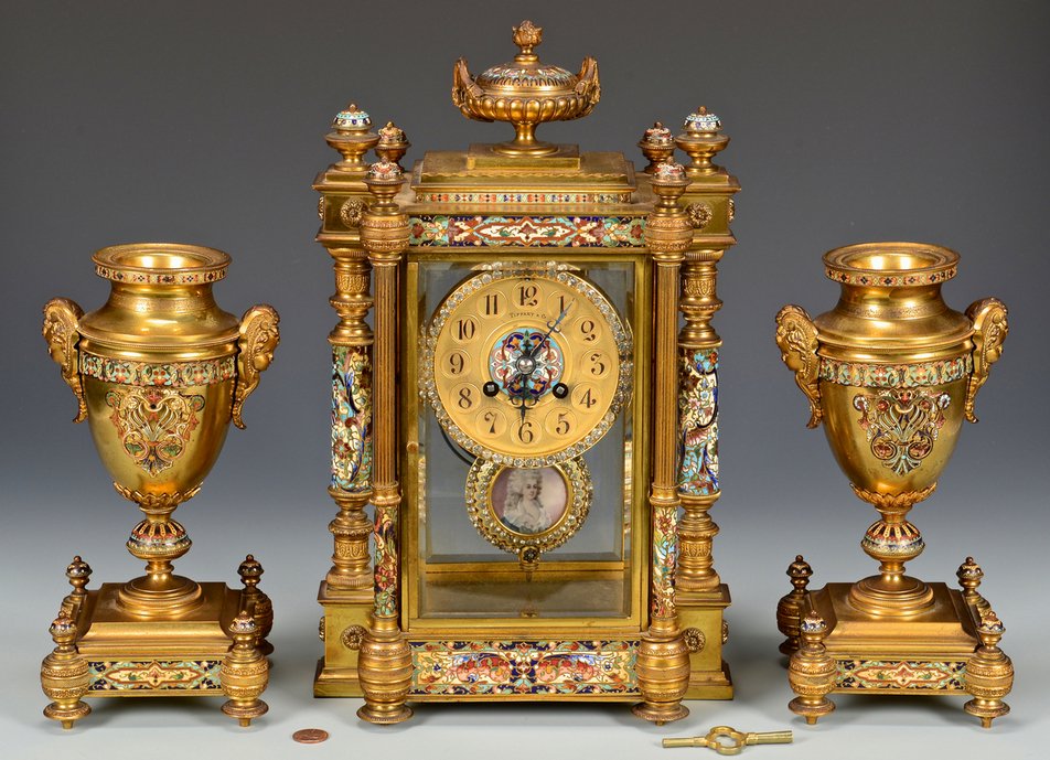 Tiffany 3-piece Garniture Set: Three-piece Tiffany Egyptian revival champleve gilt bronze garniture set with mantle clock and pair of urns. The mantle clock of gilt bronze and beveled glass with center urn finial with champleve lid