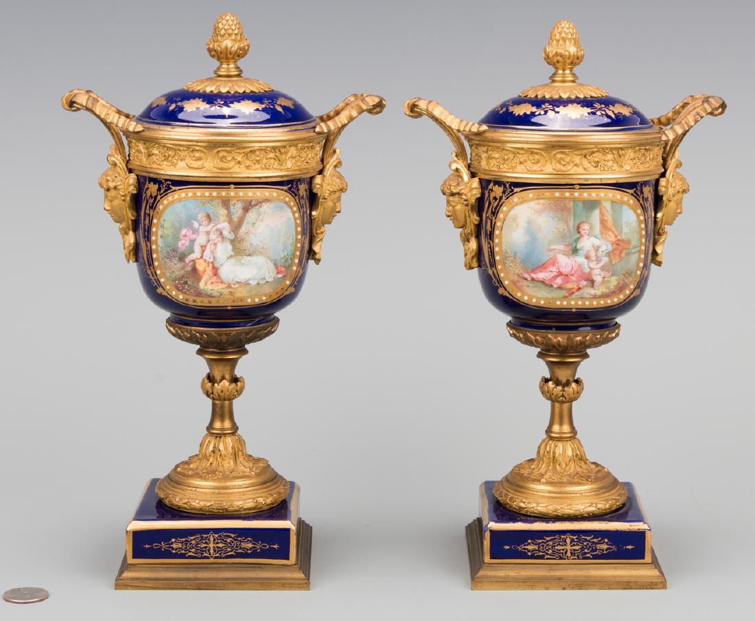Pr. Sevres Gilt Mounted Porcelain Urns: Pair of Sevres gilt mounted covered porcelain garniture urns, having cobalt grounds with hand painted vignette decoration including cupids and young women on the obverse panels, and floral sprays on t