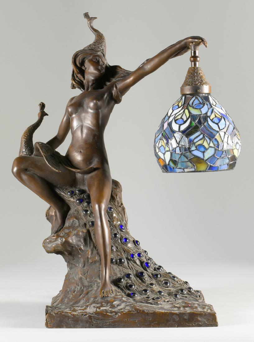 After Wolfers Bronze Nude Lamp: After Marcel Wolfers, Art Nouveau style bronze figural lamp of nude female with peacock headdress, one hand is holding a peacock with long jeweled feathers, the other hand holds a shaped stained glass