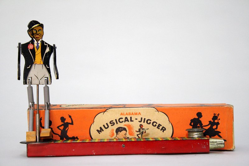 Alabama” Musical Jigger with Box: Made by Marks Bros. Co. Boston, Massachusetts –Circa 1940 This is a hand operated jigging toy with built in kazoo. As you play, he can dance. The toy is 11” long. Material: Tin
