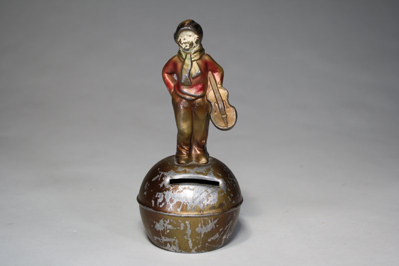 Boy with Violin on Ball Bank: Germany – Circa 1925 This is a really charming and scarce bank that we have not previously seen. He is 6” tall. Material: Spelter / Tin
