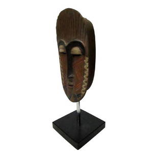 African Mask on Black Base: Oblong oval shaped African mask mounted on a black base. Mask is brown wood with tribal facial markings that could represent either face paint or traditional scarifications and a heavily stylized