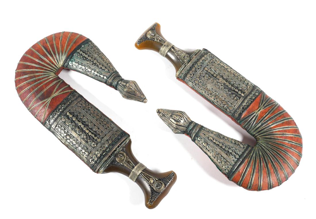 Pair of Arabian Silver Mounted Jambiya Daggers: Pair of Arabian jambiya daggers with curved double-edged blades formed with a medial ridge, silver metal mounted horn grips with leather-covered wooden scabbards. Each measures 13" long overall.