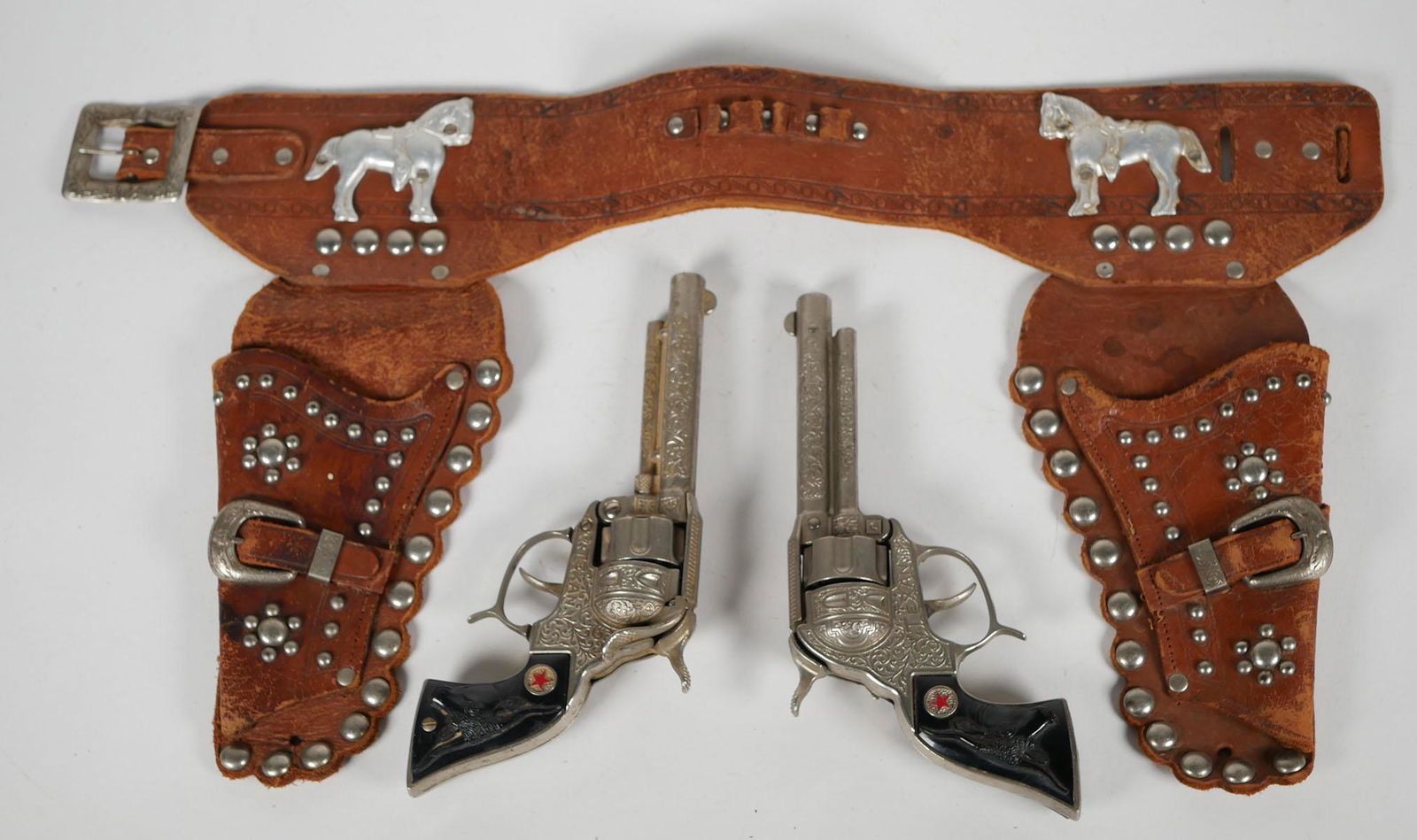 Hubley Vintage Cap Guns in Keystone Style Holster: Pair of Hubley Texan mid century red star revolvers with steers on the grips. Leather Western cowboy holster with stamped aluminum horses and round studs. Guns 9 inches long. Holster belt approx. 24 i