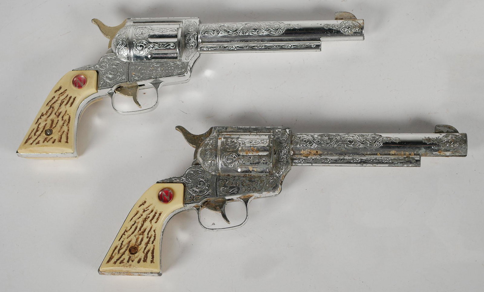 Pair of Vintage Nichols Mustang Cap Guns: Two western red star Hubley revolvers. Decorative scrollwork and faux horn ivory plastic grips. Mid century rodeo toy guns. Approx. 11 1/2 inches long. This item can be shipped in-house.