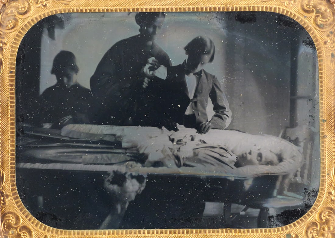 Tintype POST MORTEM Family Scene: Quarter plate post mortem. Depicting the in home viewing, the deceased is laid out in an open coffin resting on a common chair. Two young boys stand with a woman, all three mourning their loved one. A