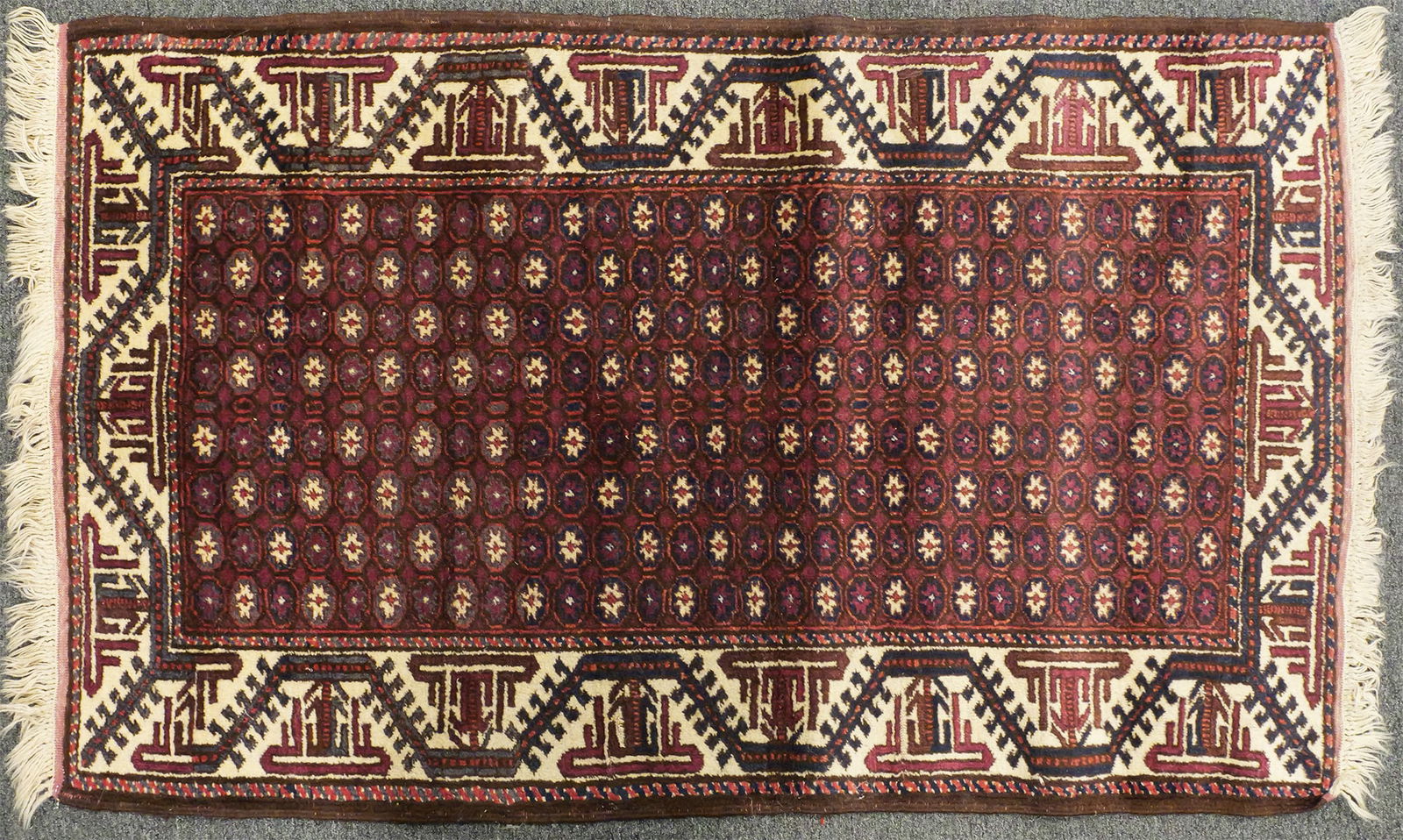 Persian Hand-Knotted Wool Rug: Hand-knotted wool rug with repeating geometric motifs on a deep red field, framed by an ivory border with angular designs. Accented with navy and rose tones and finished with fringe at both ends. Rug