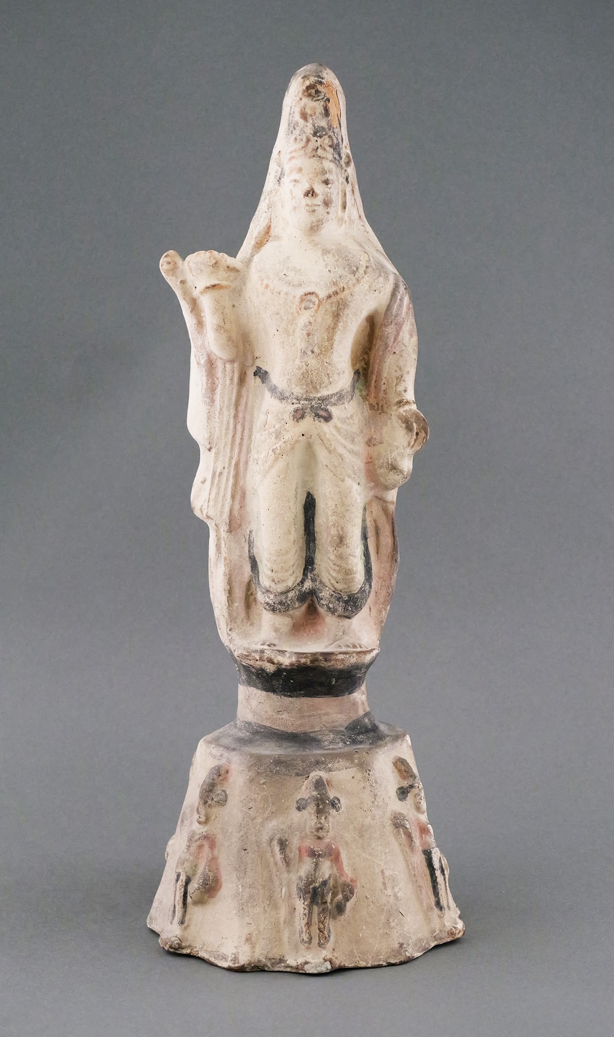 South Asian Terracotta Devotional Statue: Ancient hollow terracotta standing figure with tall headdress, mounted on integral flared pedestal with relief figures. Measures about 16-1/2" high. Traces of black over white wash. Some red shows