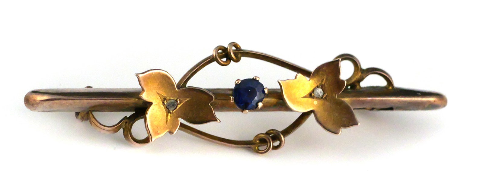 9K Gold Blue Tourmaline Floral Bar Brooch: 9K gold antique bar brooch with applied floral and scrolling decoration, set with a central round faceted blue gemstone believed to be tourmaline and two small colorless accent stones. Victorian,