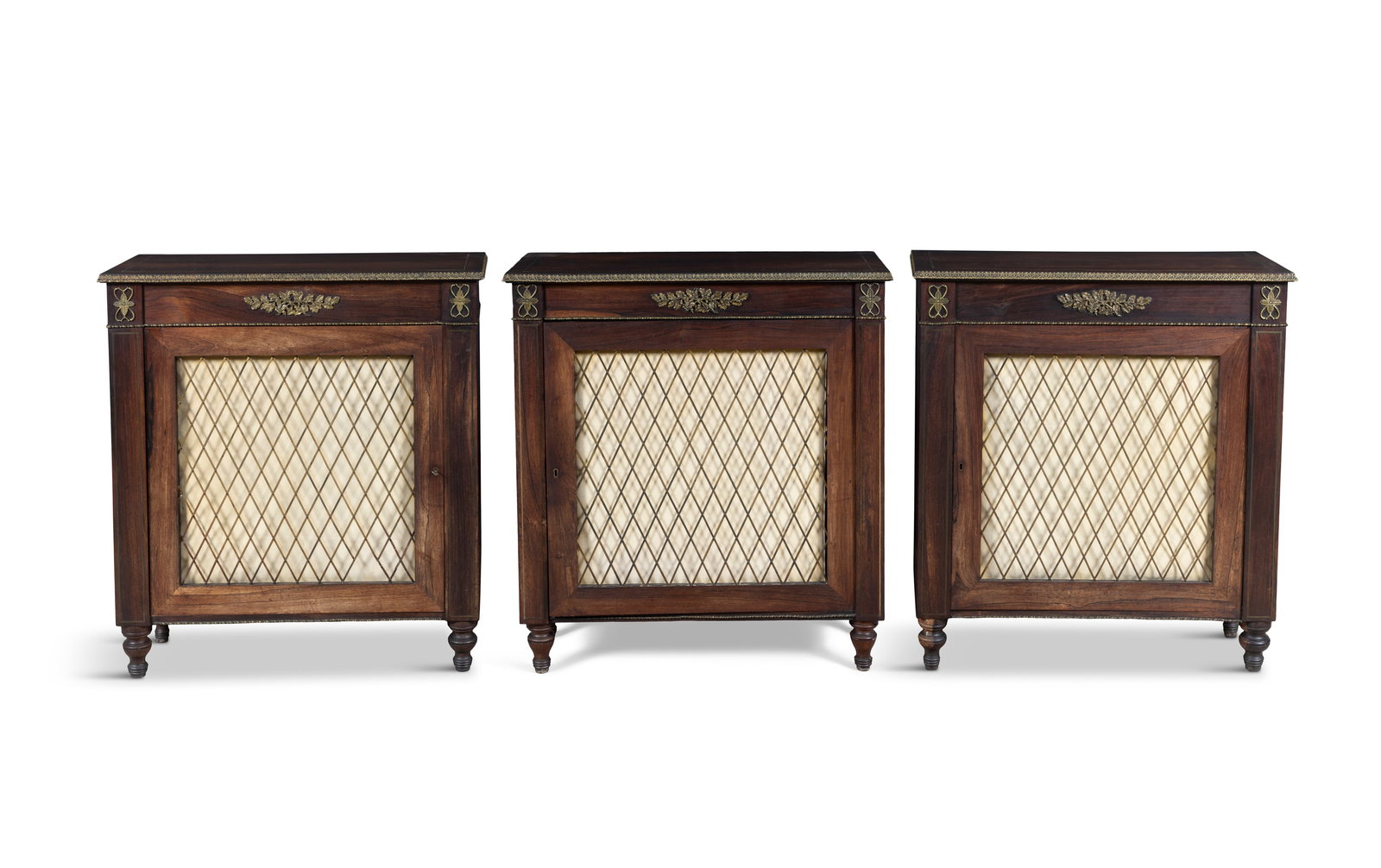 A SET OF THREE REGENCY BRASS‑MOUNTED ROSEWOOD SIDE CABINETS, c.1820, each of upright: A SET OF THREE REGENCY BRASS‑MOUNTED ROSEWOOD SIDE CABINETS, c.1820, each of upright rectangular form, the top with gilt metal trim, above a frieze centred by a cast foliate motif, flanked by an