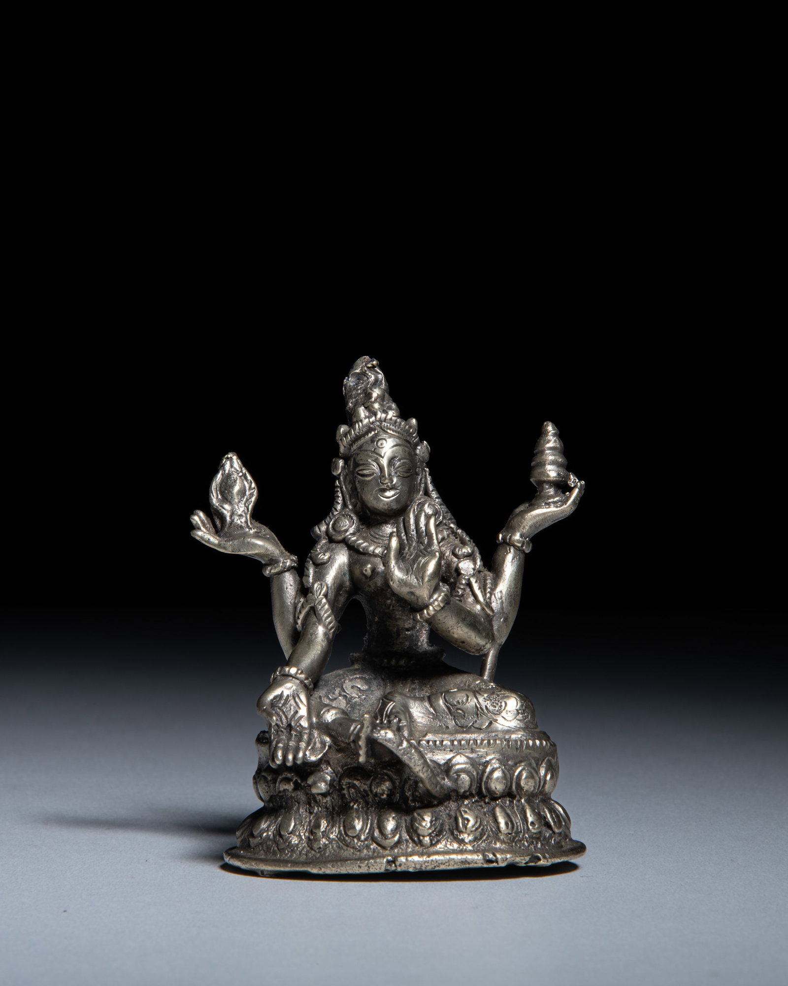 A SILVERED BRONZE ALLOY FIGURE OF A FOUR ARMS FEMININE DEITY Seated on a double-lotus base, the (1 of 3)