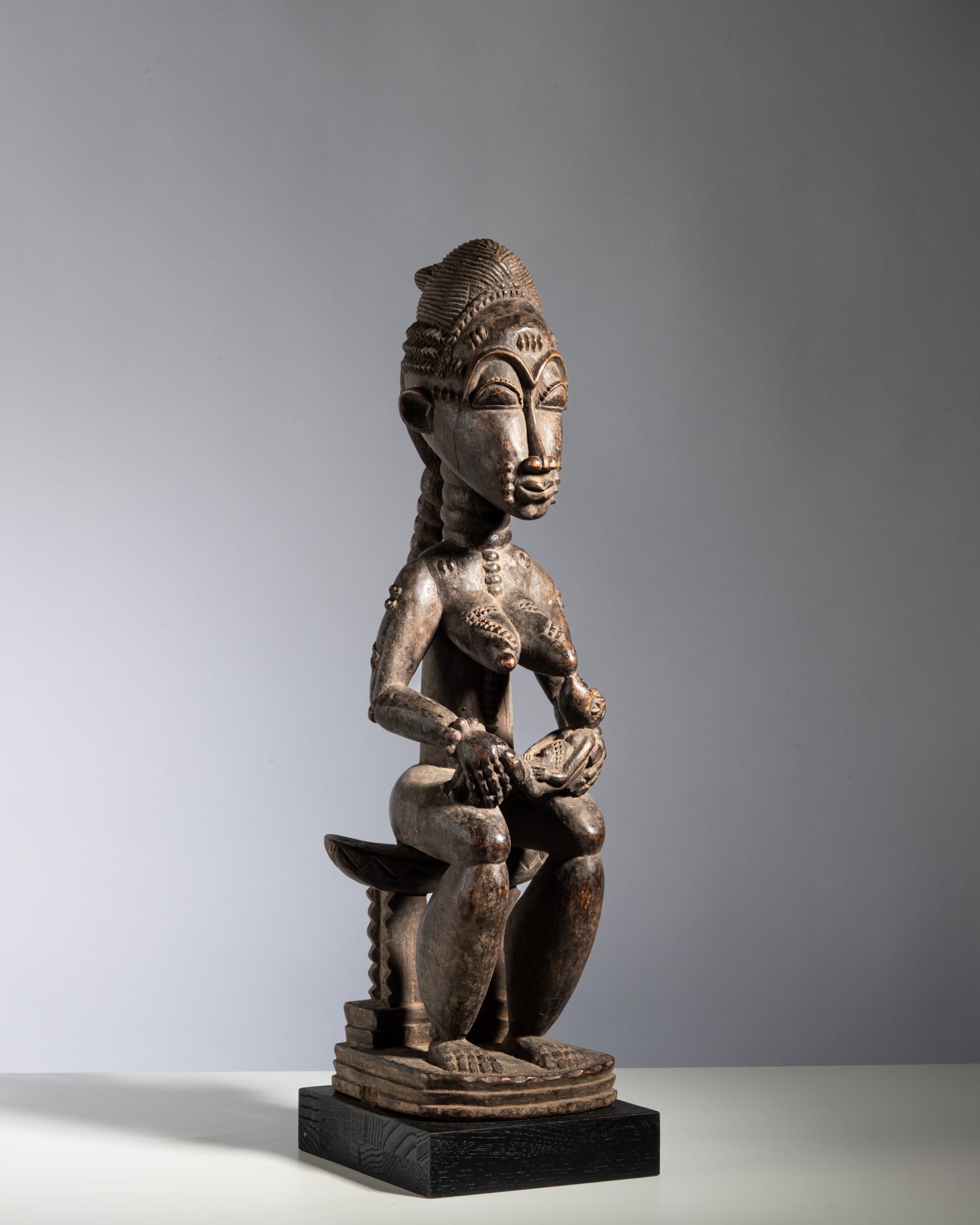 A BAOULÉ MATERNITY FIGURE Ivory Coast Wood This finely carved prestige figure depicts a: A BAOULÉ MATERNITY FIGURE Ivory Coast Wood This finely carved prestige figure depicts a seated mother nursing a child, positioned gracefully upon a traditional four-legged stool. The idealized he