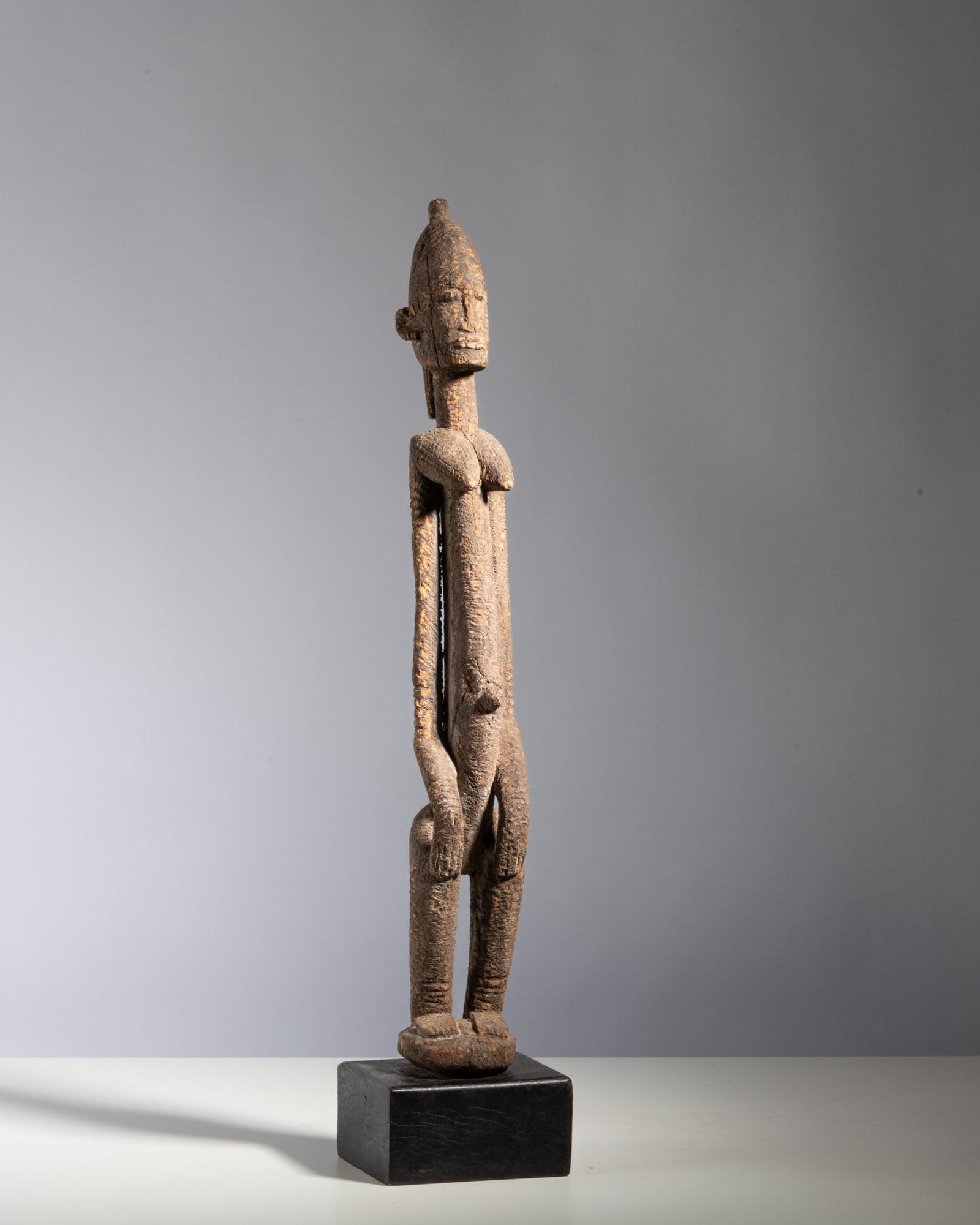 A DOGON TELLEM-STYLE FIGURE WITH RAISED ARMS Mali Wood This classic ancestral figure stands with: A DOGON TELLEM-STYLE FIGURE WITH RAISED ARMS Mali Wood This classic ancestral figure stands with exceptionally elongated, slender proportions on an integrated circular pedestal base. The figure is cap
