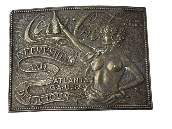 A Tiffany Studios Coca Cola Belt Buckle: A Tiffany Studios Coca Cola Belt Buckle measuring 3 5/8 inches wide by 2 3/4 inches. Made by the Tiffany foundry. Subject is a semi nude young girl holding a bottle of coke, Tiffany Foundry mark on th