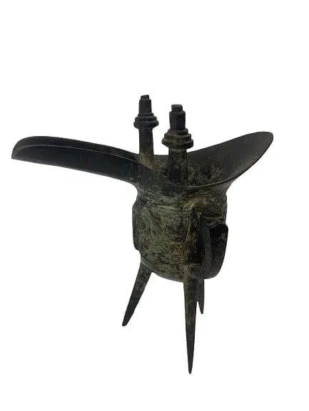 A CHINESE ARCHAIC BRONZE RITUAL WINE VESSEL (JUE) SHANG DYNASTY STYLE: A CHINESE ARCHAIC BRONZE RITUAL WINE VESSEL JUE. A bronze vessel Shang Dynasty style measuring about inches tall. deep body raised on three tall splayed knife-shaped legs, cast around the sides with s