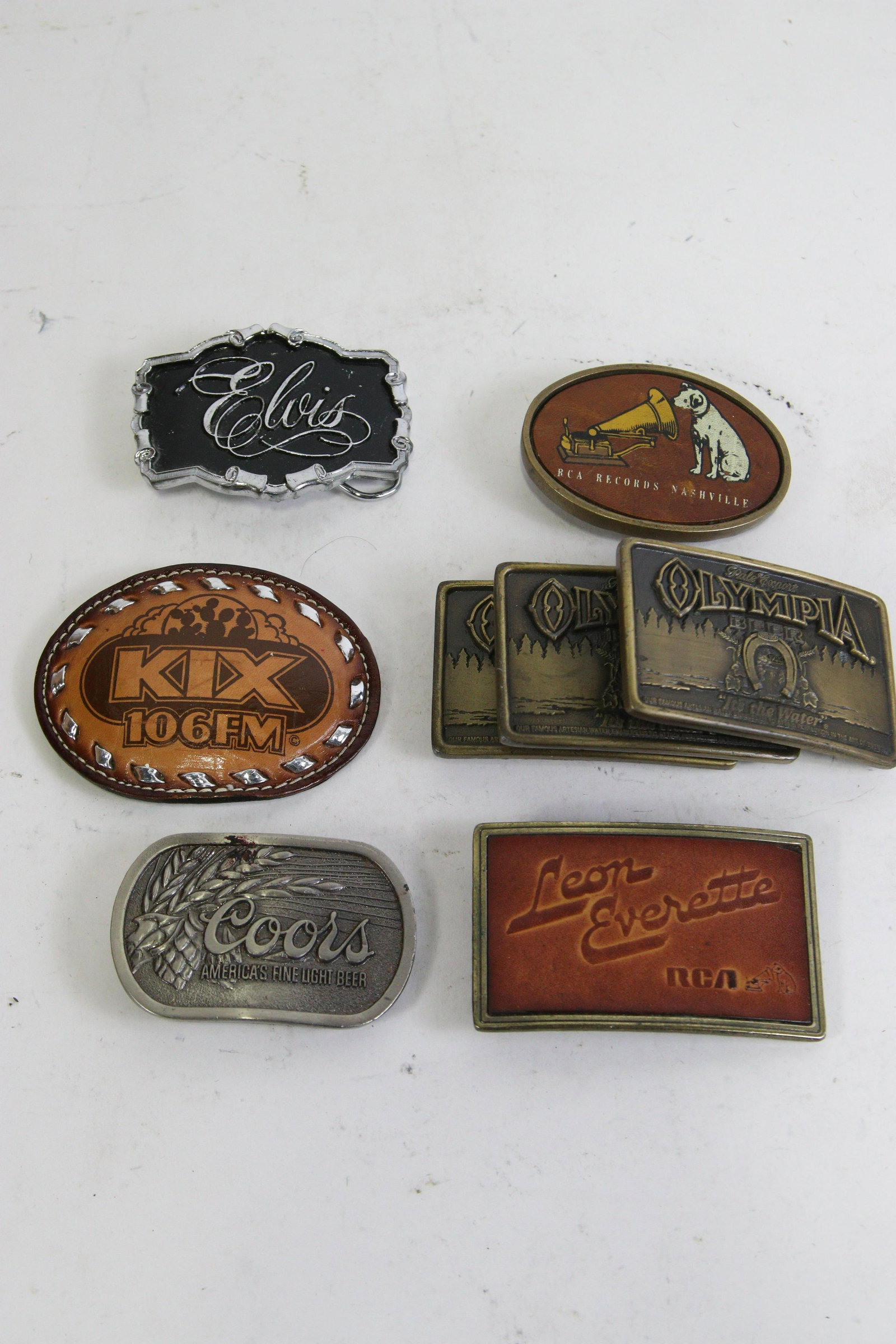 MIXED LOT OF (8) JOHN WALTON PERSONAL ITEMS: MIXED LOT OF (8) JOHN WALTON BELT BUCKLES: KIX 106, LEON EVERETT, ELVIS, COORS BEER, (3) OLYMIA BEER, & RCA RECORDS