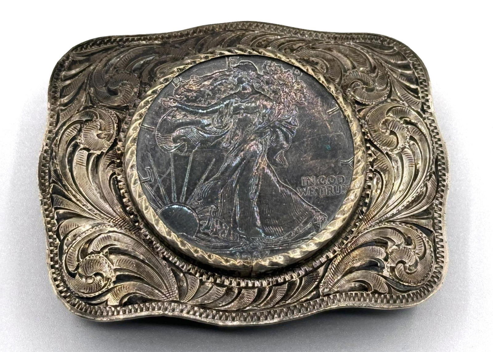 STERLING BELT BUCKLE WITH HAND CHASE WESTERN STYLE: STERLING BELT BUCKLE WITH HAND CHASE WESTERN STYLE AND US SILVER DOLLAR, STAMPED A HYKES 82.1 GRAMS Height: 2 in. by Width: 2 7/8 in. by Depth: .5 in.
