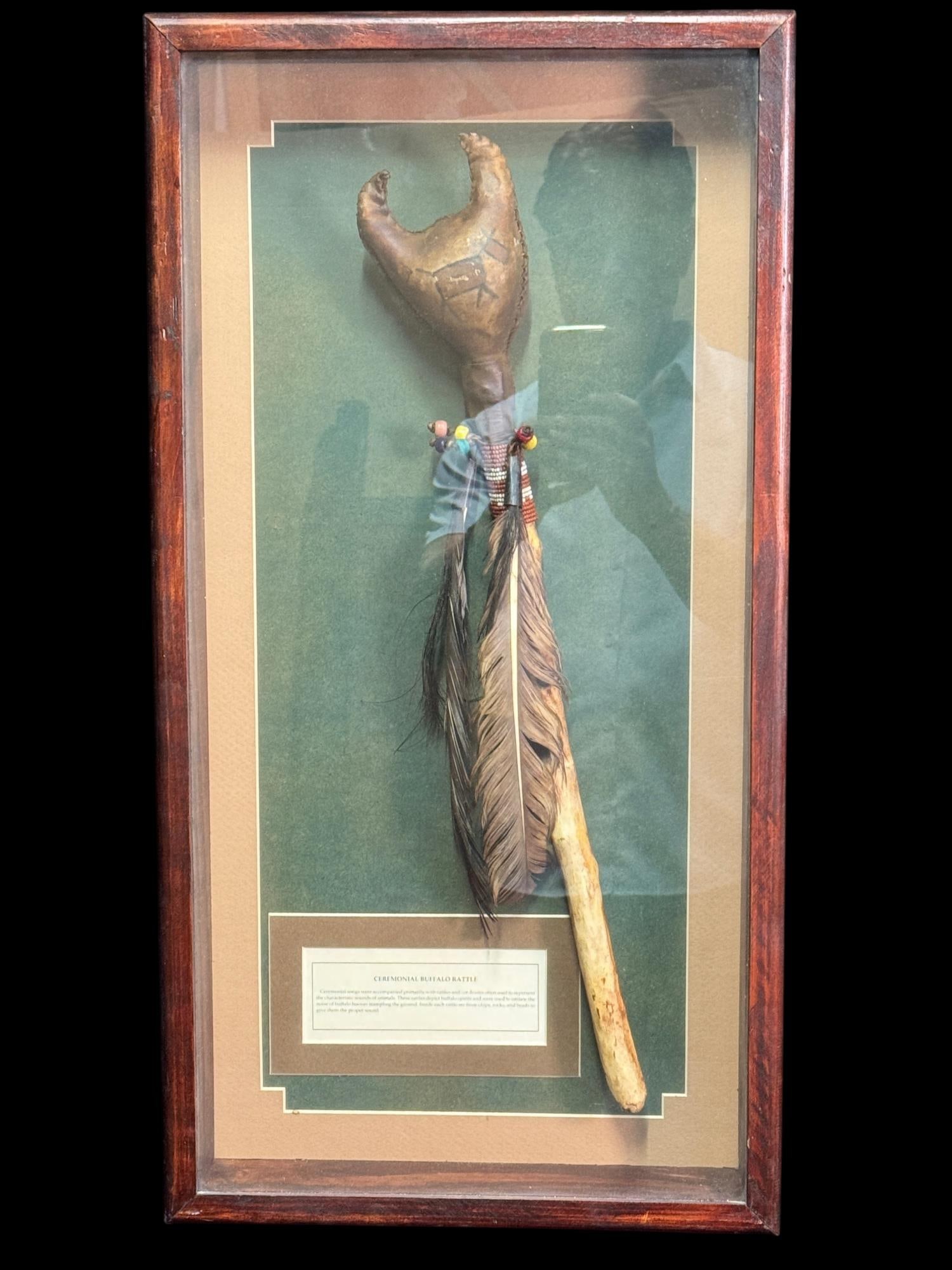 CEREMONIAL BUFFALO RATTLE-NATIVE AMERICAN: CEREMONIAL BUFFALO RATTLE. FRAMED NATIVE AMERICAN -STYLE RATTLE WITH A SCULPTED BUFFALO FORM HEAD , WOODEN HANDLE, BEADWORK, FEATHERS AND HORSEHAIR ACCENTS. MOUNTED UNDER GLASS WITH AN INFORMATIONAL