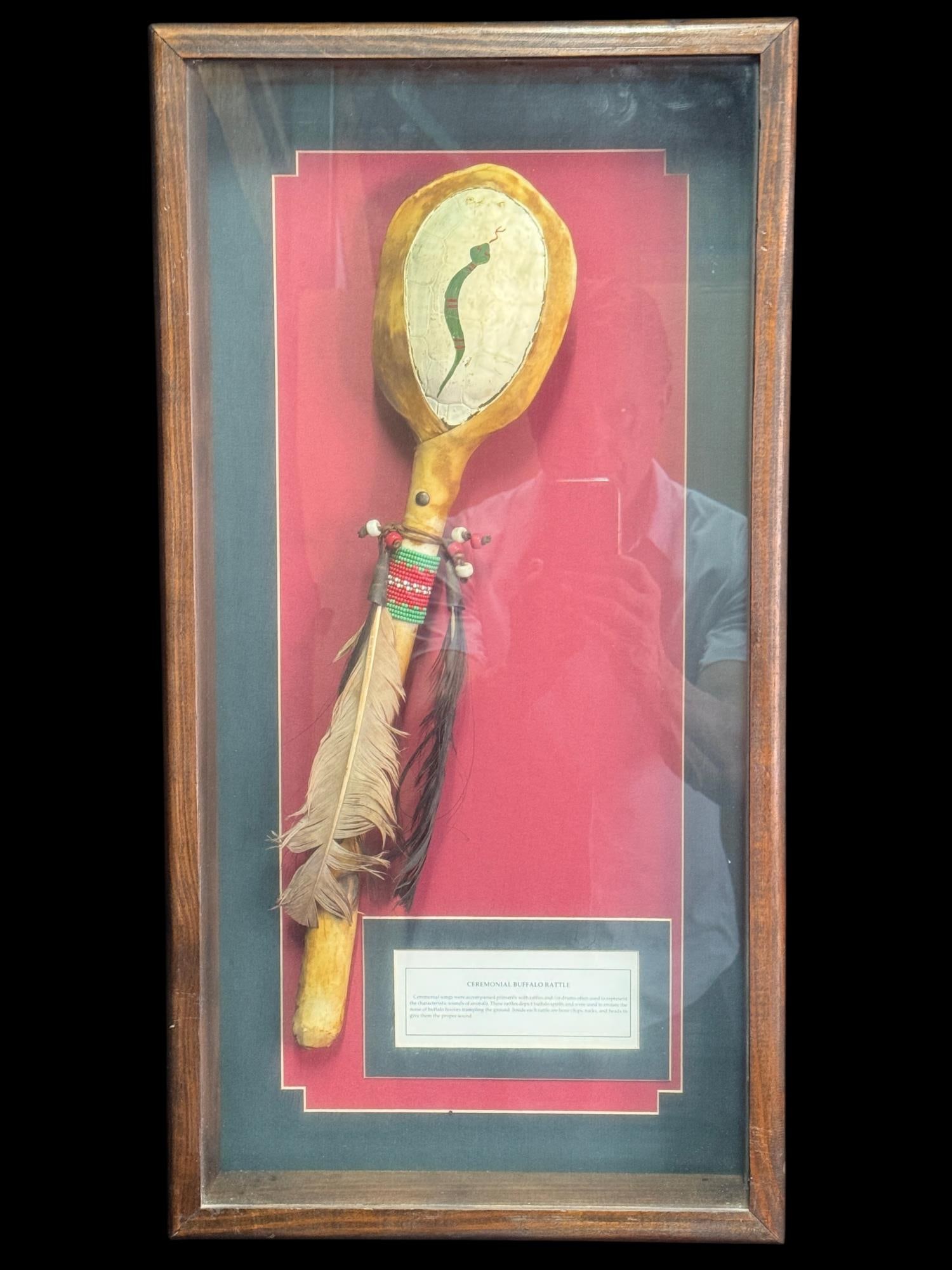 CEREMONIAL BUFFALO RATTLE. FRAMED DISPLAY: CEREMONIAL BUFFALO RATTLE. FRAMED DISPLAY WITH FEATURING A HAND DECORATED WOODEN RATTLE WITH PAINTED HIDE, BEADWORK, LEATHER AND HORSE HAIR. ACCOMPANIED BY AN INFORMATIONAL PLAQUE IN A GLAZED WOOD SHA