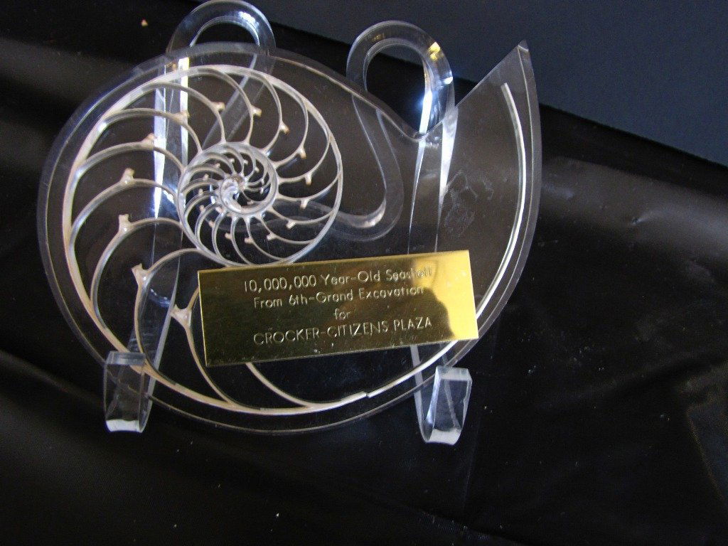 SLICE OF NAUTILUS SHELL IN LUCITE LABELLED 110T: Small slice in lucite of nautilus shell with brass label reading 10,000,000 years old from 6th grand excavation for Crocker Citizens plaza. 4.5"H x 6.25"L x 3/8"D.