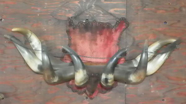 8 REAL HORN WALL COAT / HAT RACK WITH SHIELD 5014: Original hall rack of eight real horns with shield motif back in aged red velvet on wood with worn areas giving it more character, with brass chain connecting the horns on top, a great piece! C.1900.