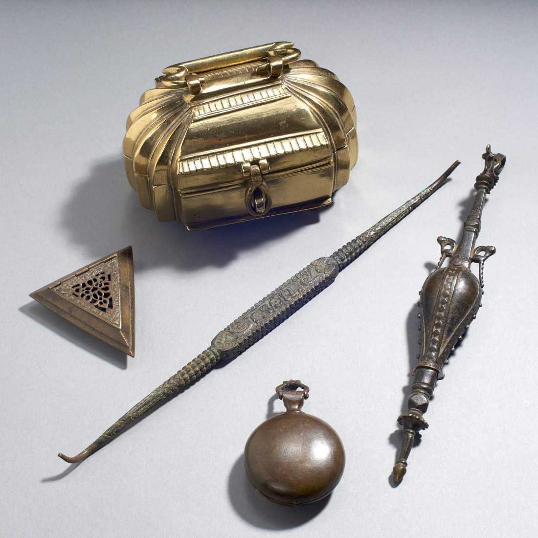 Five Indian Bronze Objects: India, ca. 18th-19th century. To include a fine 18th-century Mughal swing-handle box, an elongated woman's makeup vessel with bird finial, a tool decorated with a cold-chased design including elephant