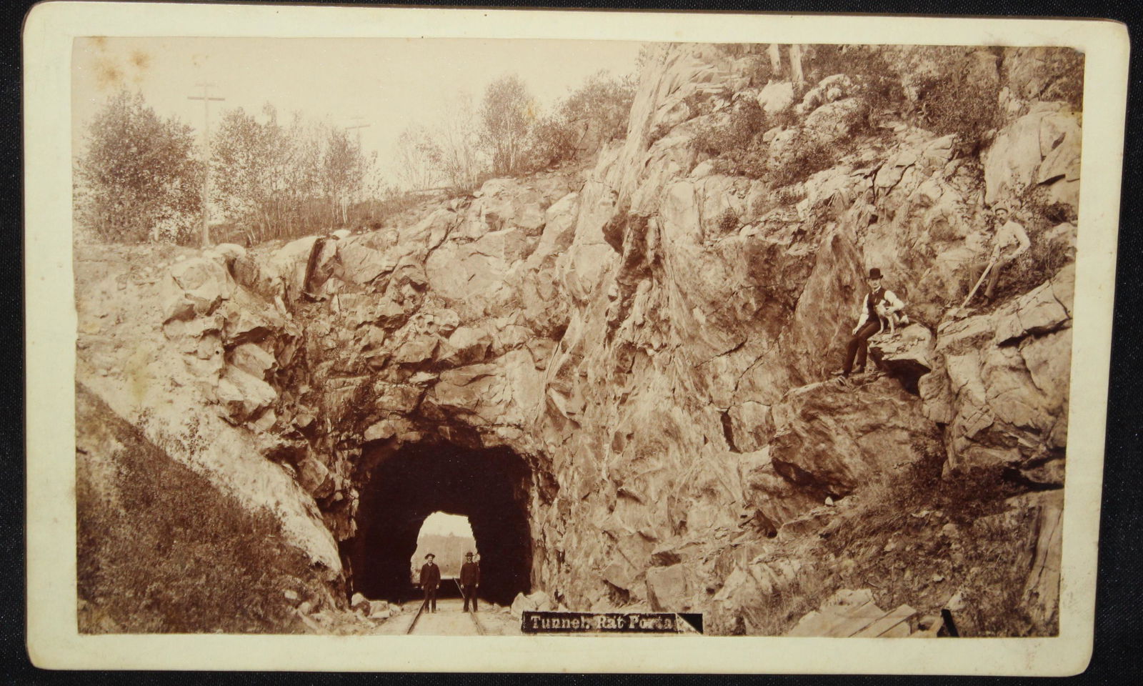 Old Photograph of Raton New Mexico Territory RR Tunnel: 5x8, circa 1890s. Photo of two men standing at the entrance of a 100ft railroad tunnel which has been blasted through of a rocky mountain ledge. Two more men and a dog are seated up on the ledge too.