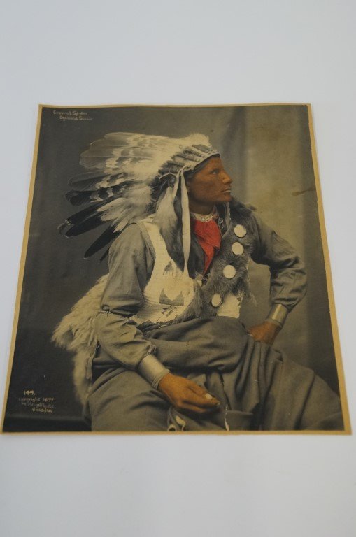 Heyn Photo - Ground Spider - Ogalla Sioux: Platinum print photo of Sioux warrior in large feathered headdress, bearskin mirroed cross-belt, red neckerchief beaded vest, wraparound blanket, dark shirt and wrist bangles. Knee-up seated view. 7x9
