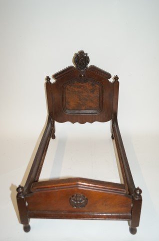 Fancy Victoration Walnut Doll's Bed: Walnut doll's bed. Crested headboard decorated with a raised Northwind god; 17.5in. H. x 22in. L. x 13in. W.