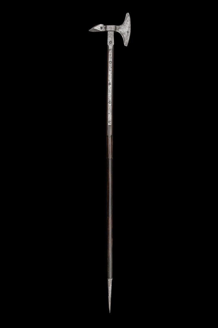 GOTHIC TOURNAMENT AXE, EARLY 16TH C.: Origin: South Germany.Steel hammered gothic tournament axe on the long, wooden shaft. The axe head has a spiky square cross section butt and semicircular blade. Maker’s hallmark hammered on the butt