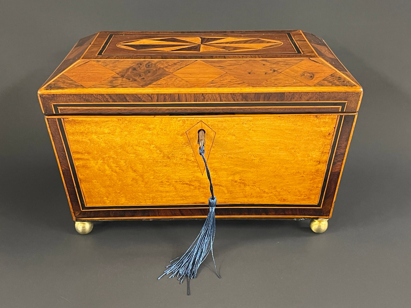 19TH C MULTI INLAID SARCOPHAGUS TEA CADDY: 19TH C MULTI INLAID SARCOPHAGUS TEA CADDY, BIRDSEYE MAPLE FON PATERAE TO TOP AND HARLEQUIN BORDER TO TOP AND HARLEQUIN LIDS TO LATCH ON BRASS BALL FEET 9.5" X 6" X 7"