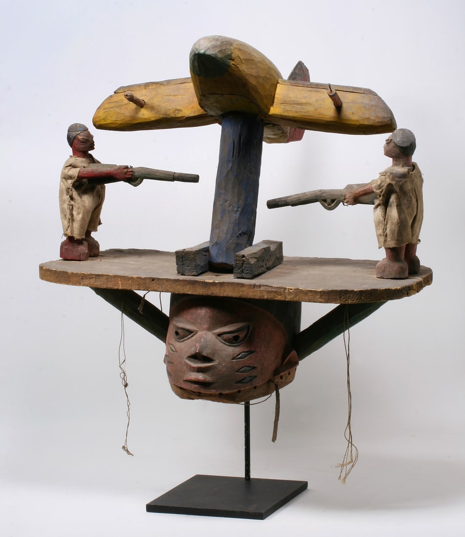 Yoruba Figural Gelede Dance Mask - Mid 20th Century: Large and heavy Gelede dance mask featuring a scene, on the top of the face and head, of two soldiers shooting down a jet war plane. Mounted on a custom made, black-painted iron display base. Carved a