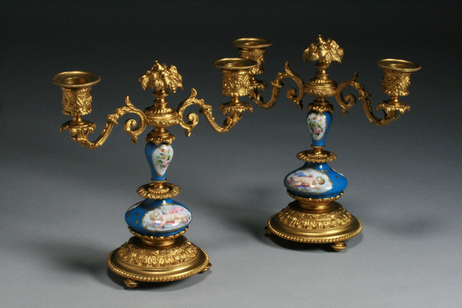 Pair of French Sevres Candlesticks - Porcelain & Gilt Bronze 19th C.: A very fine pair of French porcelain and gilt bronze candlesticks. Serves - 19th Century. Beautifully decorated with opposing Cherubs or Angels on the fronts and floral bouquet still lives on the back