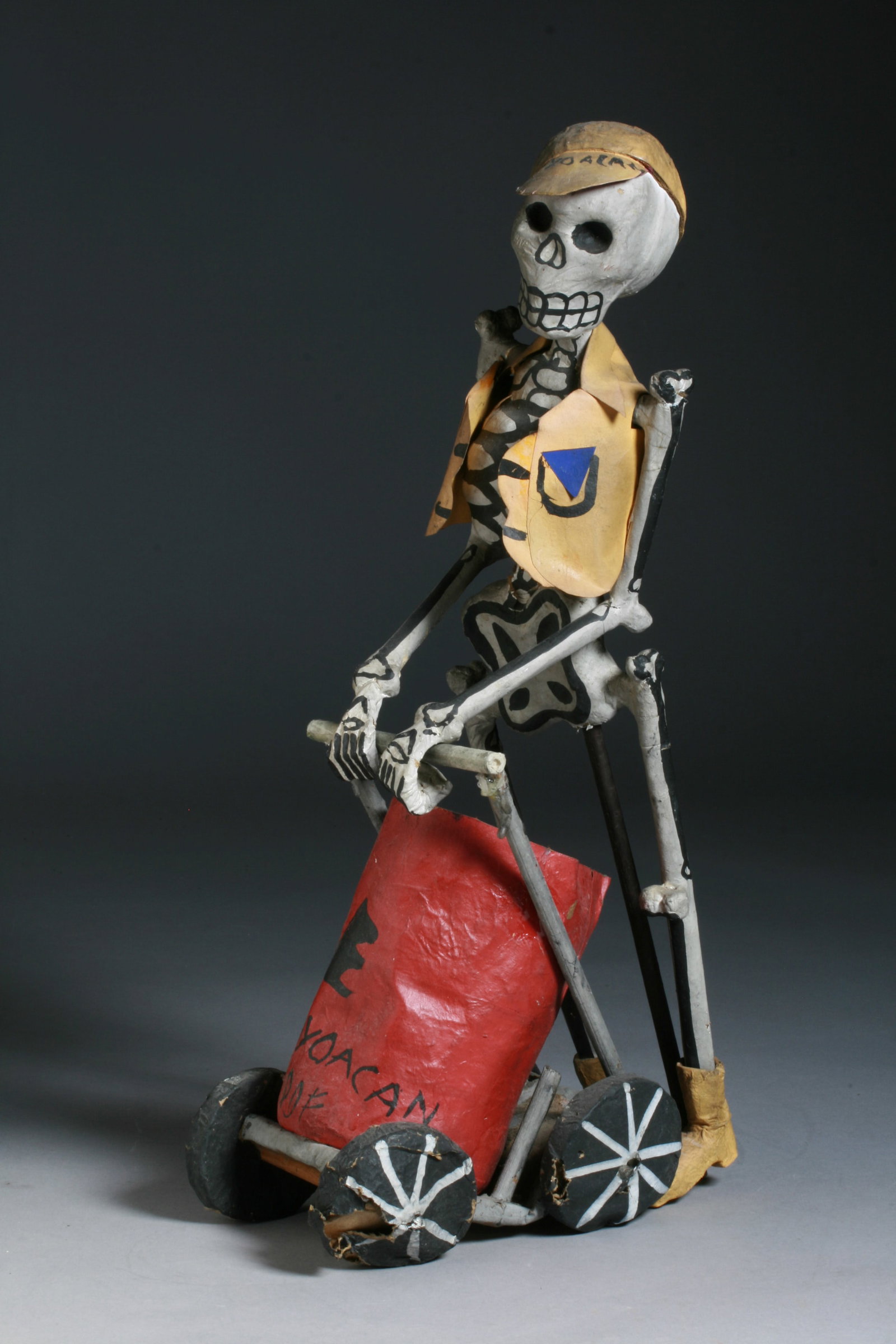 Old Mexican Day of the Dead Sculpture: A painted papier mache figural sculpture of a skeleton pushing a wheeled cart. Mid 20th century. Two pieces, the cart and the figure. A Day of the Dead figure from Coyoacan, Mexico wearing a cap and y