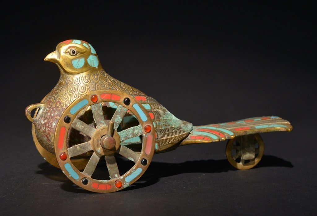 A BRONZE GEM-STONE INLAID PIGEON: HAN DYNASTY The carving is a seated pigeon on wheel. The exterior of the pigeon is decorated with colorful designs. 7 1/16 in. (18 cm) long, 3 1/2 in. (8.9 cm) tall.
