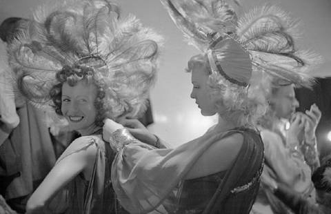 Feather Headdress: Artist: Bert Hardy Title: Feather Headdress Medium: Silver Gelatin Year: 1948 Height/Width: 16 x 20 in Description: Girls make the finishing touches to one another’s costumes before the night’s pe