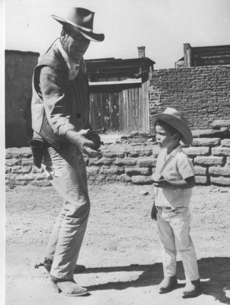 John Wayne on the set next to a child, 1965 circa: Title: John Wayne on the set next to a child, 1965 circa Dimensions: 12 x 16 Description: Agenzia Ansa. Print on polished paper. Stamp of agency on the back. In the photograph, John Wayne pulling out 