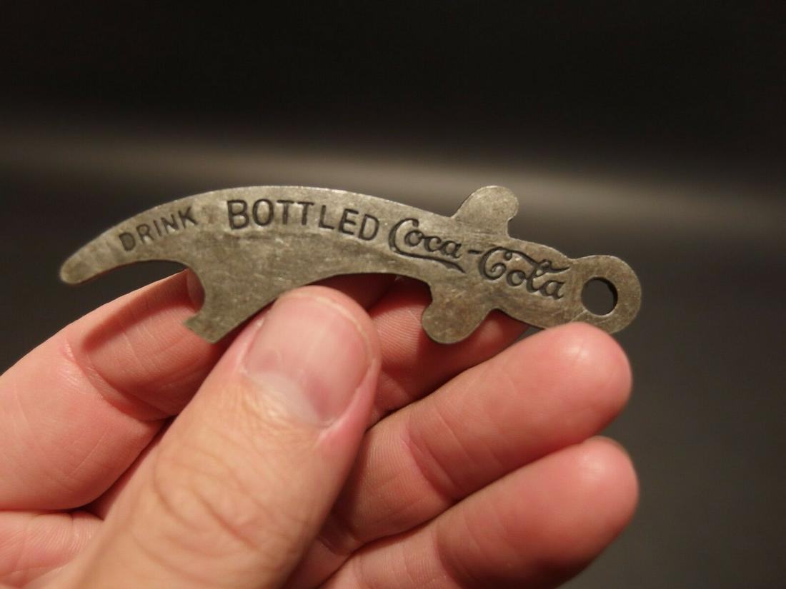 Coca Cola Bottle Opener: 3" Long Nice reproduction coca cola bottle opener shaped like a pirate sword I believe. It says Drink Bottled Coca Cola on one side then "Purity is sealed in a bottle" The opener is a nold silver pewt