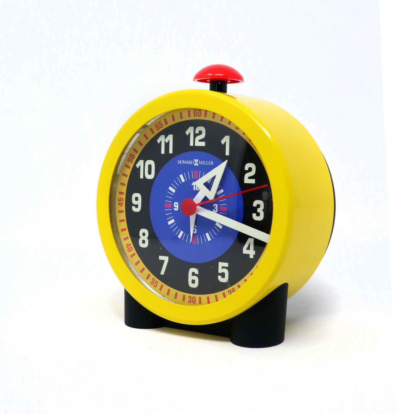 Yellow George Nelson Style Alarm Clark by Howard Miller: In the style of George Nelson's pioneering clock design for Howard Miller, this alarm clock has a multi-colored face, bright yellow metal case, and prominent red alarm button on its top. Makes a delig