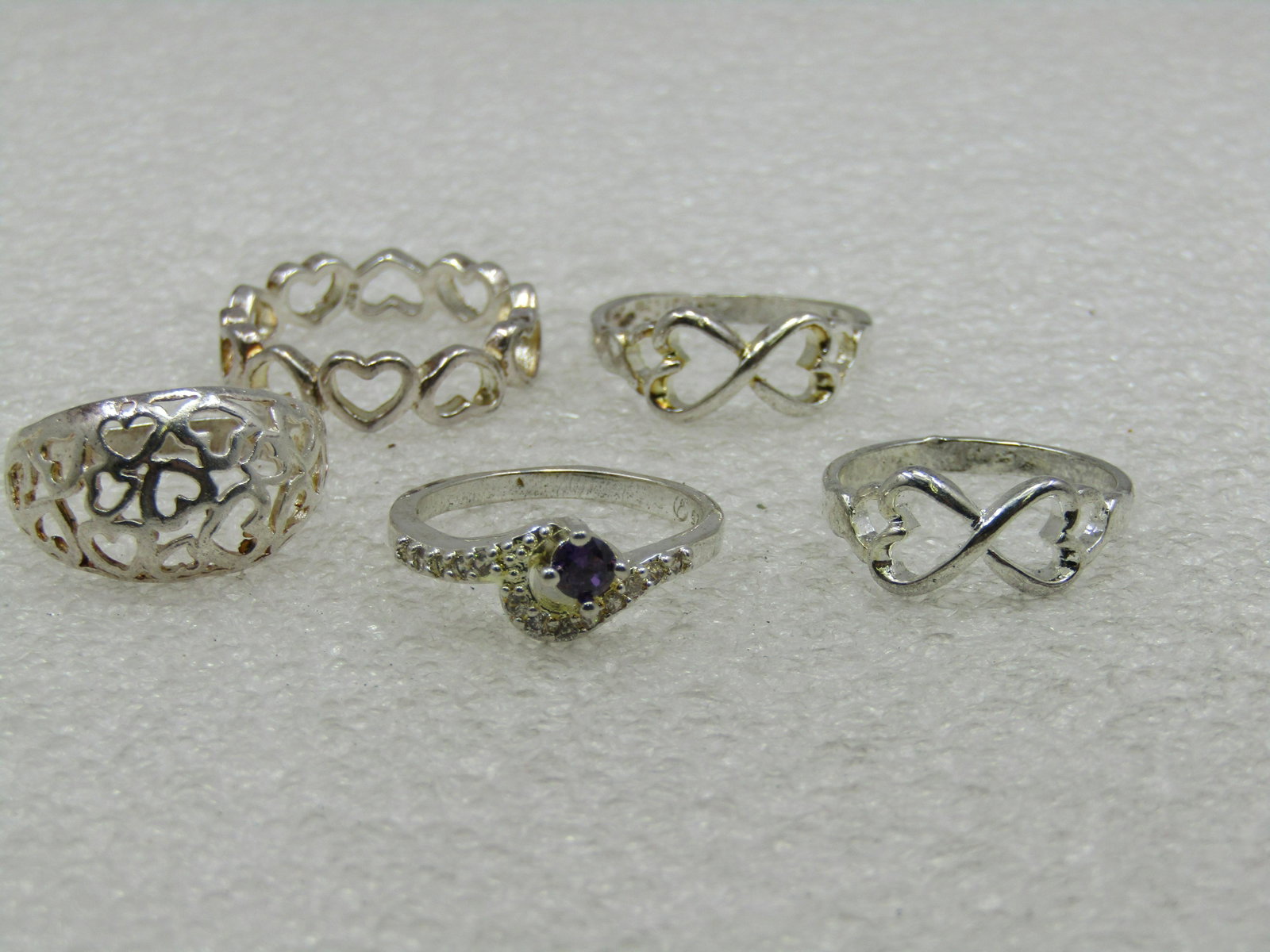 5 Silver Heart Ring Lot, Sz. 8.25 to 9, 1 with CZs: 5 Silver Heart Ring Lot, Sz. 8.25 to 9, 1 with CZs. Great lot for resale. All have a heart theme except one which has an amethyst colored center stone and clear stones around the one side of the main 