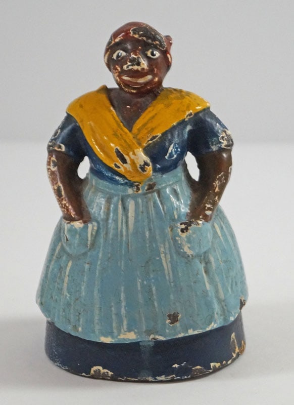 Antique Mammy Wearing Apron Cast Iron Doorstop: Title: Antique Mammy Wearing Apron Cast Iron Doorstop Dimension: 4 1/4” tall x 2 5/8” wide. Date: Ca. 1920’s. Additional Information: A full-figure, solid casting of small Mammy Anti