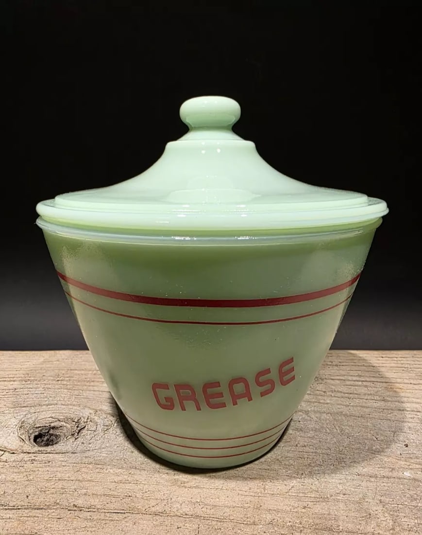 6" Jadeite Depression Glass Retro Grease Jar: 5 1/2" W x 6" H Reproduction never used jadeite green glass grease jar with a lid, often referred to as a grease drippings bowl. It features red stripes and the word "GREASE" printed on the front. The
