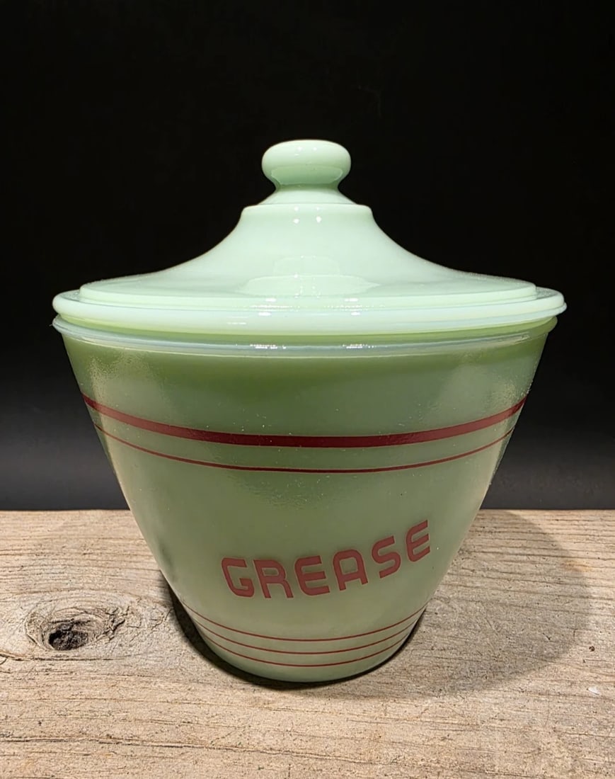 6" Jadeite Depression Glass Retro Grease Jar: 5 1/2" W x 6" H Reproduction never used jadeite green glass grease jar with a lid, often referred to as a grease drippings bowl. It features red stripes and the word "GREASE" printed on the front. The