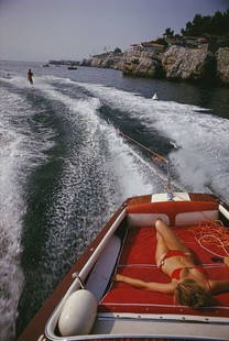 Slim Aarons 'Leisure In Antibes' 1969 C Type Print Framed In White: Description Slim Aarons 'Leisure In Antibes' 1969 C Type Print Framed In White A woman sunbathing in a Riva motorboat as it tows a waterskiier, in the sea off the Hotel du Cap-Eden-Roc in Antibes on t